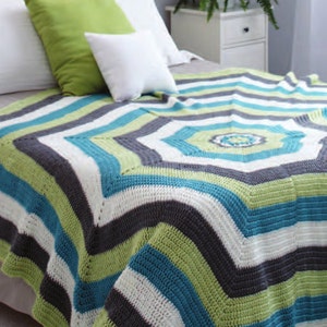 May include: A striped crocheted throw blanket in shades of green, white, gray, and blue. The blanket is folded on a bed with pillows.