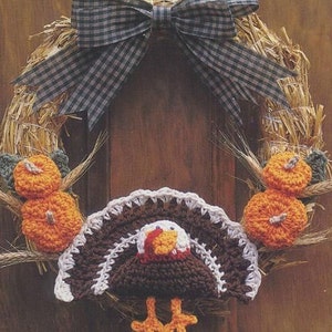 May include: A crocheted Thanksgiving wreath featuring a brown turkey, orange pumpkins, and a black and white checked bow. The wreath is hanging on a wooden door with the text "Happy Thanksgiving" written below.