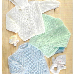 Puede incluir: Tres jerséis de punto para bebé en blanco, verde claro y azul claro con patrones de punto de cable. Los jerséis están colocados sobre una superficie de madera. El texto "DOUBLE KNITTING" está en la parte inferior de la imagen.