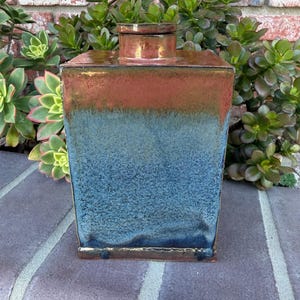 May include: A rectangular ceramic canister with a lid, featuring a gradient glaze that transitions from a deep, textured blue at the base to a warm, metallic copper-toned finish at the top, with a smooth, flat-topped cylindrical neck and lid.