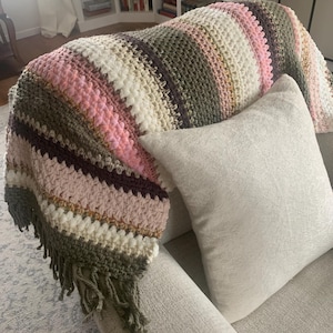 May include: A multi-colored crocheted throw blanket with fringe on a light gray couch. The blanket has stripes of pink, white, brown, and green.