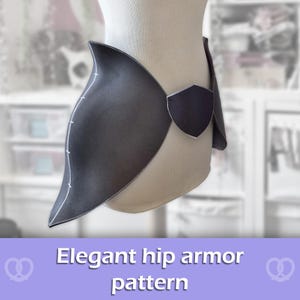 May include: A dark gray hip armor piece featuring a curved, flared silhouette with a central shield-shaped accent. The edges are defined by a light gray trim, and the surface displays a smooth, matte finish with subtle white line markings.