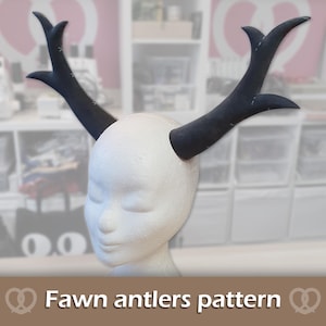 May include: A white foam mannequin head wearing a pair of black, branched antlers. The antlers feature visible white stitching along the edges. Below the head, a brown banner displays the white text Fawn antlers pattern with pretzel icons.