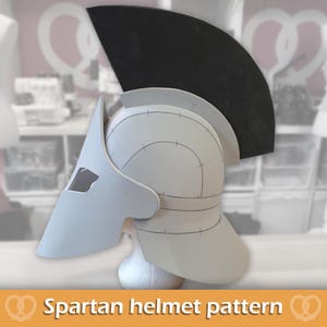May include: A light gray Spartan helmet pattern constructed from foam or cardstock, featuring a prominent black crest. The helmet displays visible assembly lines and markings on its surface, with a distinct nose guard and neck protection piece.