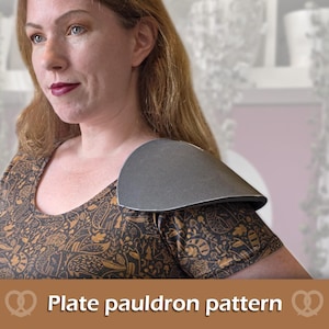 May include: A dark gray, curved plate pauldron resting on the shoulder. The pauldron has a smooth, matte finish and a rounded, protective shape. Below the image, the text reads: Plate pauldron pattern, flanked by two pretzel icons.