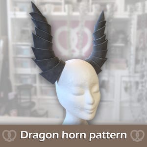 May include: A pair of dark grey, spiralled dragon horns displayed on a white mannequin head. The horns feature a layered, segmented construction that tapers to a point at the top. The text "Dragon horn pattern" is printed at the bottom of the image.