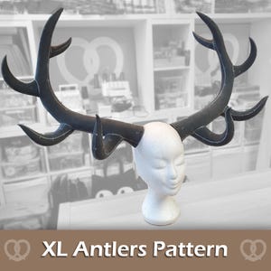 Puede incluir: Patrón de cuernos XL. Un par de cuernos grandes de color gris oscuro se muestran en una cabeza de maniquí blanca. Los cuernos tienen múltiples puntas curvas y están cosidos con un hilo más claro. El texto "XL Antlers Pattern" está en la parte inferior.