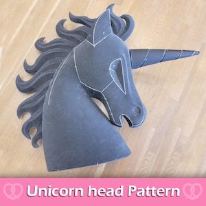 Può includere: Un modello di testa di unicorno grigio scuro con linee di cucitura bianche, un corno a spirale e una criniera fluente. Il testo "Unicorn head Pattern" è visualizzato in rosa e bianco nella parte inferiore dell'immagine. La testa è appoggiata su una superficie di legno marrone chiaro.