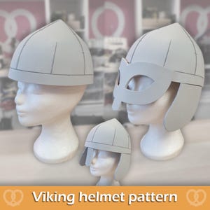 Può includere: Tre modelli di elmo vichingo in schiuma grigio chiaro esposti su teste di manichino bianche. I modelli presentano una costruzione a cupola segmentata, con un design che include maschera e alette laterali, e un altro con alette laterali e cuciture.