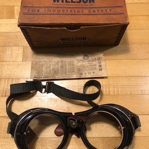 May include: Vintage Willson safety goggles with a black frame and clear lenses, accompanied by their original brown box and paperwork. The box reads "FOR INDUSTRIAL SAFETY." The goggles have a black strap.