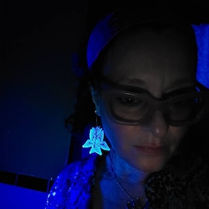 May include: A woman wearing a blue patterned shirt and glasses with glowing blue butterfly earrings.
