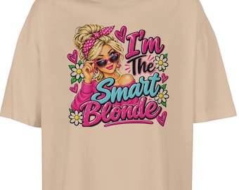 May include: A beige t-shirt featuring a graphic of a blonde person with a pink polka-dot headband and sunglasses. The text reads I'm The Smart Blonde in pink and blue lettering, surrounded by white daisies and pink hearts.