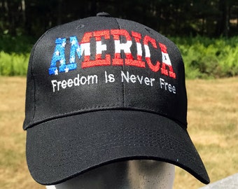 May include: Black baseball cap with the word "AMERICA" embroidered in red, white, and blue. The text "Freedom Is Never Free" is embroidered in white below the word "AMERICA".