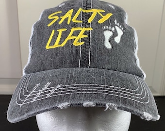 May include: A black and white distressed baseball cap with a yellow embroidered design that says "Salty Life" and white footprints.