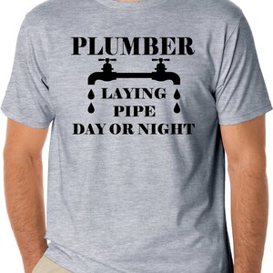 May include: A gray t-shirt with a black graphic of two faucets with water drops and the text "Plumber Laying Pipe Day or Night".