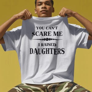 Może przedstawiać: Biała koszulka z czarnym napisem "YOU CAN'T SCARE ME I RAISED DAUGHTERS". Koszulka jest trzymana przez osobę. Tło jest w jednolitym ż&oacute;łtym kolorze.