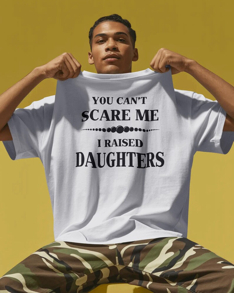 Może przedstawiać: Biała koszulka z czarnym napisem "YOU CAN'T SCARE ME I RAISED DAUGHTERS". Koszulka jest trzymana przez osobę. Tło jest w jednolitym ż&oacute;łtym kolorze.