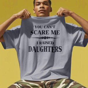 Może przedstawiać: Jasnoszara koszulka z czarnym napisem "YOU CAN'T SCARE ME I RAISED DAUGHTERS". Koszulka jest trzymana przez osobę. Tło jest ż&oacute;łte.