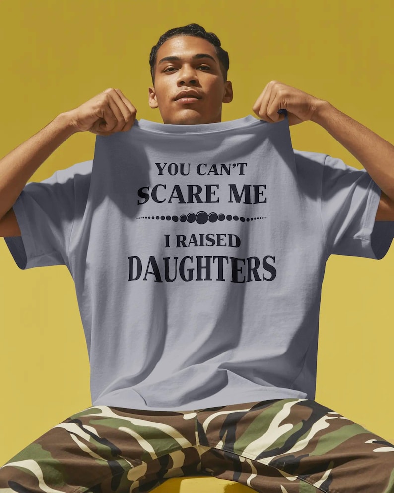 Może przedstawiać: Jasnoszara koszulka z czarnym napisem "YOU CAN'T SCARE ME I RAISED DAUGHTERS". Koszulka jest trzymana przez osobę. Tło jest ż&oacute;łte.
