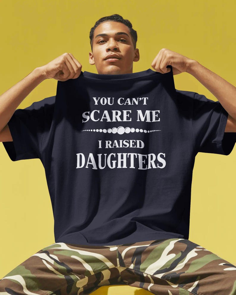 Może przedstawiać: Granatowa koszulka z białym napisem "YOU CAN'T SCARE ME I RAISED DAUGHTERS". Koszulka jest trzymana przez osobę. Tło jest ż&oacute;łte.