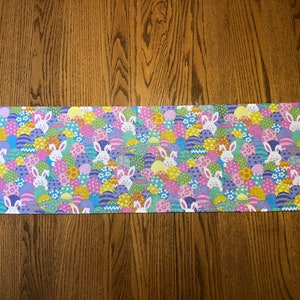 May include: A purple and green table runner with a pattern of Easter eggs and bunnies. The eggs are in shades of blue, yellow, pink, and green.