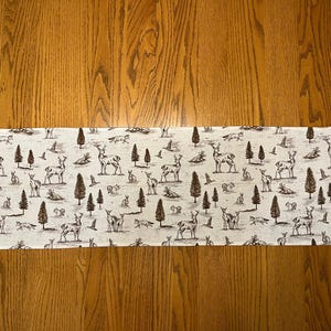 May include: A rectangular table runner with a repeating pattern of deer, rabbits, birds, and trees in brown and white. The runner is placed on a wooden table, showcasing a woodland theme.
