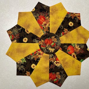 May include: A decorative, circular fabric piece with a fall theme. The design features alternating sections of solid mustard yellow and patterned brown fabric. The brown fabric is decorated with pumpkins, leaves, and flowers. The center has a pumpkin and leaf design.