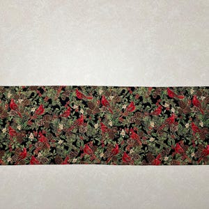 May include: A decorative table runner featuring a repeating pattern of red cardinals, pine cones, and greenery on a black background. The runner has a pointed end design and is made of fabric.