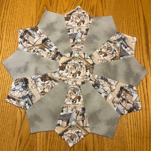 May include: A star-shaped Christmas table topper with alternating sections. The gray sections are solid, while the other sections feature a print of a nativity scene in shades of brown, beige, and white. The topper is placed on a wooden surface.