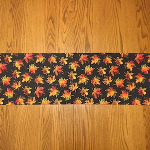 May include: A black table runner with a pattern of colorful fall leaves. The leaves are in shades of orange, yellow, and red.