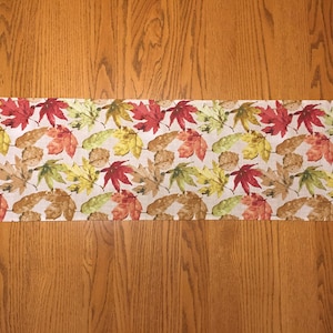 May include: A beige table runner with a pattern of red, yellow, and green leaves. The runner is 14 inches wide and has a pointed end.