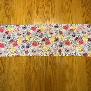 May include: A white table runner with a floral pattern featuring pink, purple, and yellow flowers on a white background.