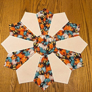 May include: A decorative, star-shaped table topper with a fall theme. The design features alternating sections of beige fabric and fabric printed with pumpkins in shades of orange, teal, and white. The topper is likely used for seasonal home decor.
