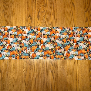 May include: A table runner with a fall theme featuring a pattern of orange, white, and blue pumpkins.