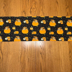 May include: A black table runner with a repeating pattern of orange pumpkins with cats peeking out of the top. The pumpkins have green vines and white polka dots on a black background.