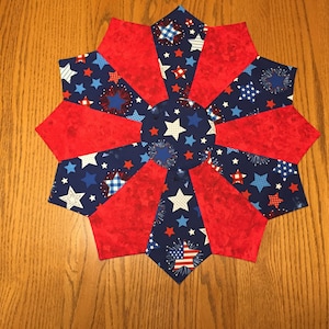 May include: A patriotic, star-shaped table topper with a red, white, and blue color scheme. The design features alternating red panels and blue panels with white stars and fireworks. The center is a blue circle with white stars.