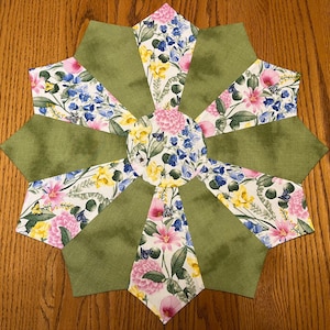 May include: A green and white floral table runner with a starburst design. The runner features a central circle with a floral pattern and is surrounded by 12 green points.