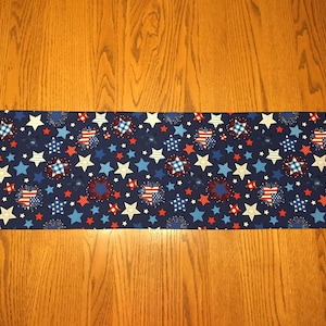 May include: A blue table runner with a patriotic design featuring red, white, and blue stars and fireworks. The runner has a pointed end on each side.