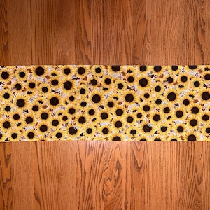 May include: A rectangular table runner with pointed ends featuring a dense, repeating pattern of yellow sunflowers with dark brown centers and small green leaves on a light background, displayed flat against a wood grain surface.
