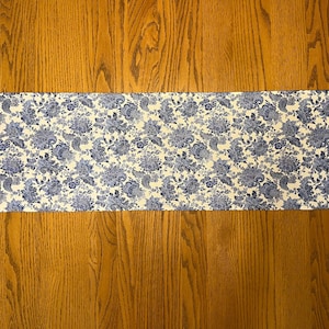 Puede incluir: Camino de mesa rectangular con un estampado floral en tonos azules y blancos. El camino de mesa está colocado sobre una superficie de madera. El diseño floral presenta flores y follaje detallados, creando un aspecto clásico y elegante. El camino de mesa mide unos 30 cm de ancho.