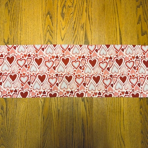 May include: A white table runner with a pattern of red and pink hearts. The runner is on a wooden table.