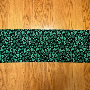 May include: A black table runner with a green shamrock pattern. The runner is approximately 14 inches wide and 72 inches long.