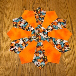 May include: A round orange and blue fabric table runner with a pumpkin pattern. The runner is made of multiple fabric pieces sewn together to create a starburst design.