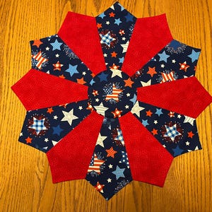 May include: A star-shaped quilted table topper with a patriotic theme. The design features alternating red fabric segments and navy blue segments with white stars, fireworks, and American flag patterns. The center is navy blue with a white star.