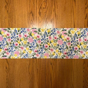 May include: A white table runner with a floral pattern featuring pink, yellow, and blue flowers. The runner has a pointed end on each side.