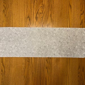 May include: A light gray table runner with a repeating pattern of white stars and swirls. The runner is rectangular with angled ends. The fabric is laid on a wooden surface.