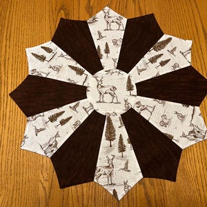 May include: A star-shaped table runner with a woodland theme. The design features alternating dark brown and patterned segments. The patterned sections display deer, rabbits, trees, and birds in shades of brown and white.
