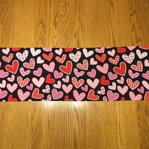May include: A black table runner with a pattern of red and pink hearts. The hearts are of various sizes and have different patterns, including polka dots, stripes, and gingham.