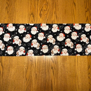May include: A black table runner with a repeating pattern of Santa Claus faces. The Santa Claus faces are white with red hats and white beards. The background is black with a brick pattern and white stars.
