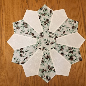 May include: A white and green table runner with a pine cone and branch pattern. The runner is made of fabric and has a sparkly finish.
